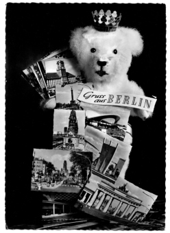 Postkarte mit Berliner Bär und Mauerkrone und dem Schriftzug „Gruss aus BERLIN“. Der Bär hält diverse Postkarten aus Berlin vor seinem Körper